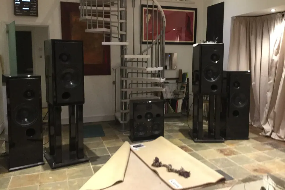 Several pairs of tall floor-standing loudspeakers on stands in a living room with a tiled floor, a rolled-up rug and a spiral staircase behind